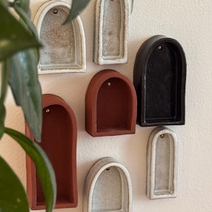Op de afbeelding: Een verzameling kleine, gewelfde keramische wandplanken in verschillende kleuren, waaronder wit, grijs, rood en zwart. Elke plank heeft een klein messingkleurig accent aan de bovenkant. De planken zijn aan een lichtgekleurde muur bevestigd.