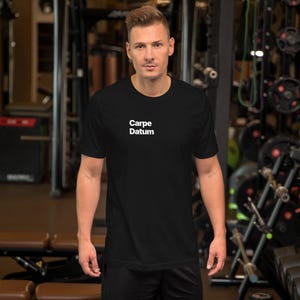 May include: Black t-shirt with the white text "Carpe Datum" centered on the chest. The shirt is short-sleeved and appears to be made of a soft material. The background is a gym setting with exercise equipment.