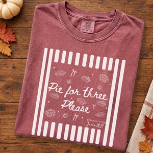 May include: A maroon crew neck t-shirt featuring a white graphic of vertical stripes, various pie illustrations, pacifiers, and bows. The text reads "Pie for three Please" and "twins arriving June 2027". The shirt has a Comfort Colors brand label.