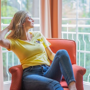 Könnte beinhalten: Ein gelbes T-Shirt mit einem kleinen schwarzen und blauen Design auf der Brust. Die Person trägt eine blaue Jeans und sitzt in einem roten Stuhl. Der Hintergrund umfasst ein Fenster mit weißen Rahmen und orangefarbenen Vorhängen.