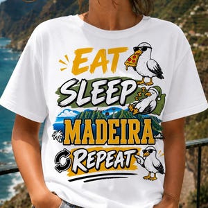 Puede incluir: Una camiseta blanca de manga corta con un estampado gráfico con el texto EAT SLEEP MADEIRA REPEAT. El diseño incluye tres ilustraciones de gaviotas, una comiendo una porción de pizza, una durmiendo y una señalando, junto a una escena de paisaje.