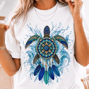 May include: A white t-shirt featuring a graphic of a sea turtle with a mandala-patterned shell in shades of blue and teal. The turtle is surrounded by a splash of water and is adorned with hanging blue and teal feathers and a small crescent moon.