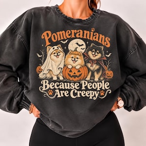 May include: A charcoal gray crewneck sweatshirt featuring a graphic of three Pomeranian dogs in Halloween costumes. The text reads "Pomeranians Because People Are Creepy" with illustrations of a ghost, a pumpkin, a vampire cape, and bats.