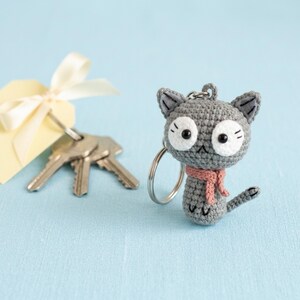 May include: A gray crochet cat keychain with large white eyes, a pink scarf, and a silver keyring. The cat has a small tail and is next to a set of keys and a yellow tag with a white ribbon.