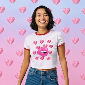May include: A white ringer t-shirt with red trim on the collar and sleeves. The front features a graphic of bright pink lips biting a lower lip, surrounded by a pattern of small pink hearts. The shirt is paired with blue denim jeans.