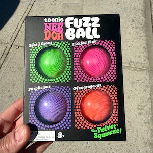May include: A black box featuring four colorful Fuzz Balls in Zesty Green, Tickled Pink, Purplicious, and Orangrageous. The box reads "teenie FUZZ DOH BALL" and "The Velvet Squeeze!" with a warning for children under 3 years old.