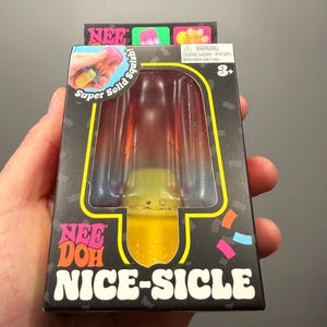 Op de afbeelding: Een Nee Doh Nice-Sicle speelgoed in een doos. Het speelgoed heeft de vorm van een ijslolly met een doorzichtige plastic buitenkant en een kleurrijk, squishy interieur. De doos is zwart met de woorden "Nee Doh" en "Nice-Sicle" in wit en oranje.