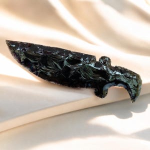 Puede incluir: Un cuchillo de obsidiana tallado a mano con una hoja afilada y un mango curvado. El cuchillo es de un negro profundo y brillante, que refleja la luz. La hoja tiene una superficie texturizada.