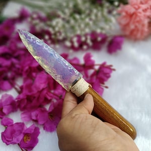 May include: A handcrafted knife with a translucent, iridescent blade and a wooden handle. The blade is secured to the handle with a wrapped, natural-colored cord. The knife is displayed against a backdrop of pink and white flowers.