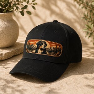 May include: A black baseball cap featuring a rectangular patch on the front. The patch displays a detailed illustration of a Bernese Mountain Dog set against a backdrop of pine trees, jagged mountain peaks, and a large sun in warm orange tones.