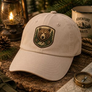 May include: A beige baseball cap featuring a shield-shaped patch on the front. The patch displays a brown bear head surrounded by green pine trees, pine cones, and mountain peaks. The cap has a structured crown, a curved brim, and stitched eyelets.