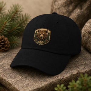 May include: Black baseball cap with a brown bear emblem. The emblem is a shield shape with a bear's face, pine trees, and mountains. The cap is made of a sturdy material and has a curved peak. A fashionable accessory for outdoor activities.