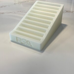 May include: A white, stepped display stand with the letters "PSA" embossed on the front. The stand has multiple horizontal slots, resembling a staircase. The object is made of a solid, opaque material.