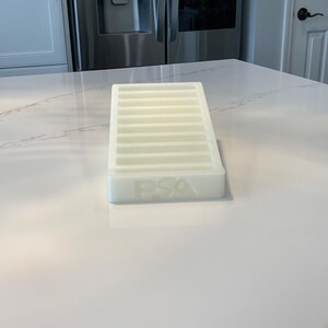 May include: A white, rectangular tray with multiple parallel slots. The tray has the letters "FSA" embossed on the front. The tray is sitting on a white countertop.