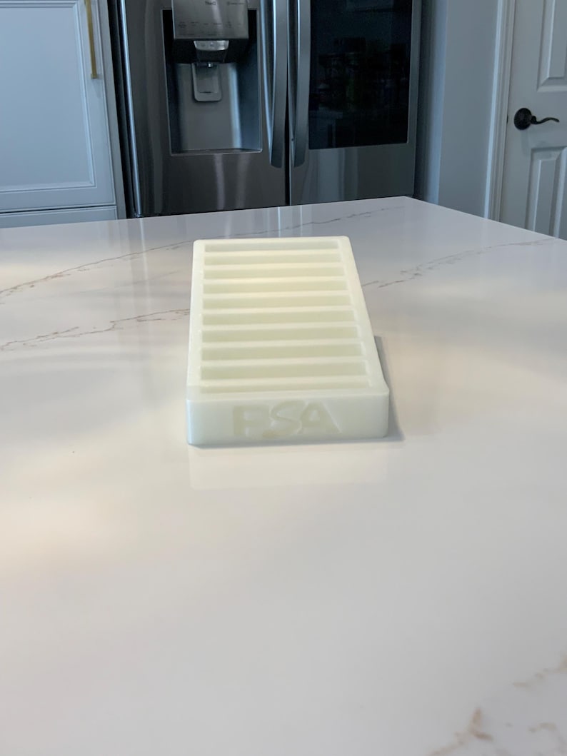May include: A white, rectangular tray with multiple parallel slots. The tray has the letters "FSA" embossed on the front. The tray is sitting on a white countertop.