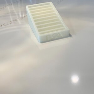 May include: A white, rectangular soap dish with a sloped design and horizontal ridges. The letters "PSA" are embossed on the front. The dish is made of a translucent material and sits on a white countertop.