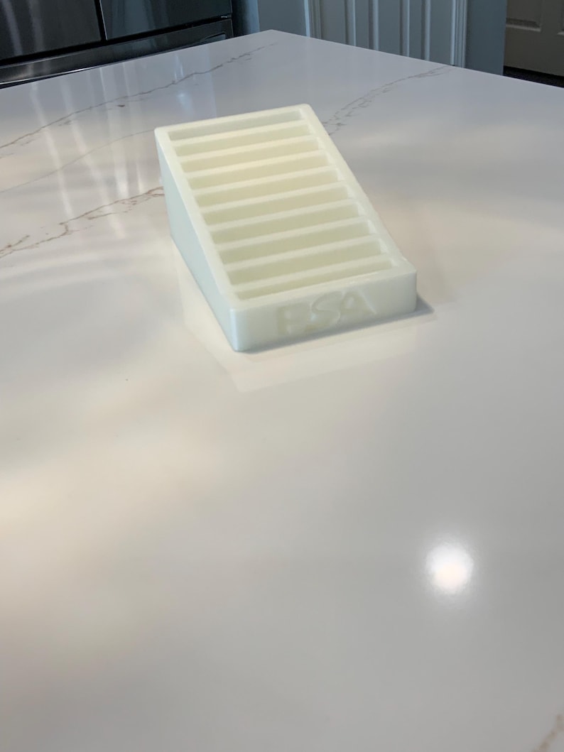 May include: A white, rectangular soap dish with a sloped design and horizontal ridges. The letters "PSA" are embossed on the front. The dish is made of a translucent material and sits on a white countertop.