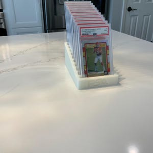 May include: A white, tiered display stand holding multiple graded sports cards. The cards are in clear protective cases, with red and gold accents visible. The text "2017 PANINI DONRUSS OPTIC PATRICK MAHOMES" is visible on one card. The stand has the letters "PSA" on the front.