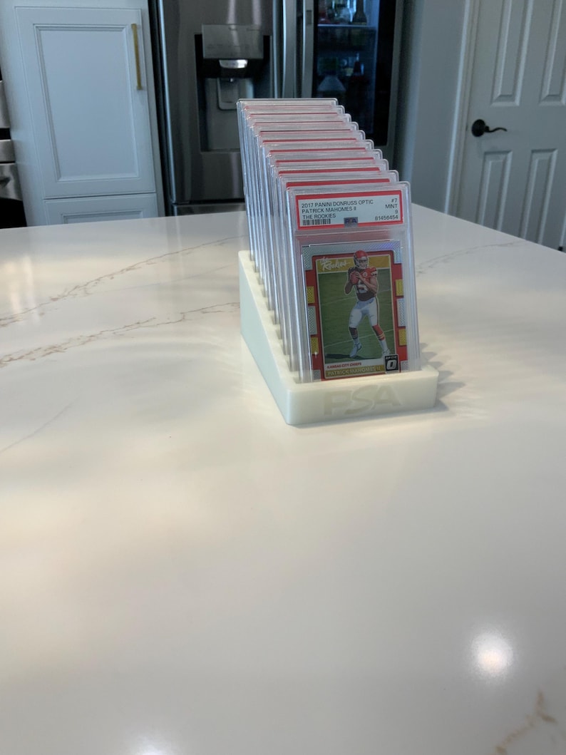 May include: A white, tiered display stand holding multiple graded sports cards. The cards are in clear protective cases, with red and gold accents visible. The text "2017 PANINI DONRUSS OPTIC PATRICK MAHOMES" is visible on one card. The stand has the letters "PSA" on the front.