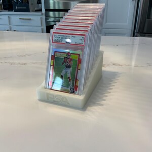 May include: A collection of graded sports cards in protective cases, displayed in a white holder. The cards feature a football player in a red uniform. The holder is labeled with the letters "PSA".