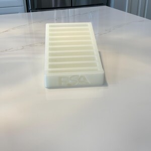 May include: A rectangular, off-white ice cube tray with multiple horizontal dividers. The letters "FSA" are embossed on the side. The tray is made of a translucent material and sits on a white countertop.