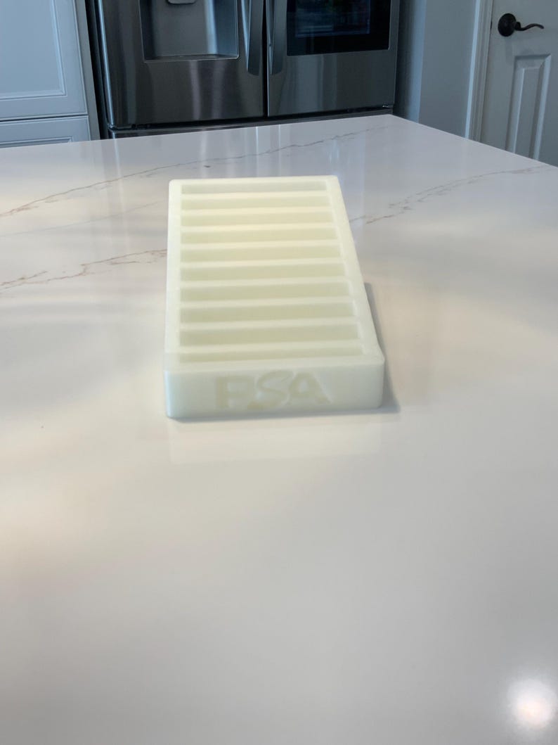 May include: A rectangular, off-white ice cube tray with multiple horizontal dividers. The letters "FSA" are embossed on the side. The tray is made of a translucent material and sits on a white countertop.