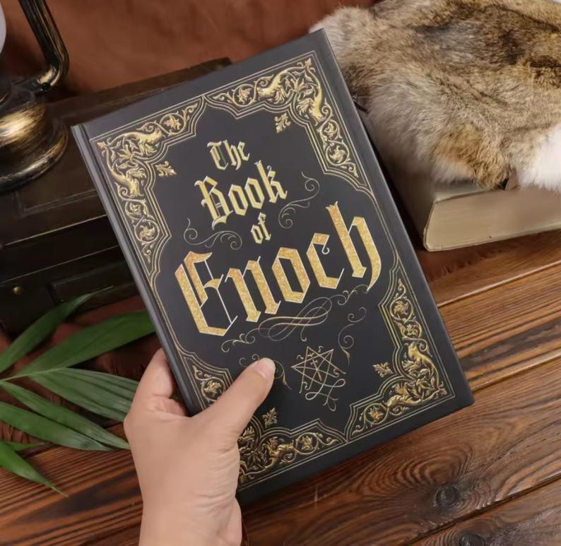 May include: A black hardcover book titled The Book of Enoch with gold foil lettering and intricate gold filigree border designs on the front cover. The cover features a central geometric symbol and ornate corner flourishes in a metallic gold finish.