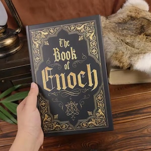 May include: A black hardcover book titled The Book of Enoch with gold foil lettering and intricate gold filigree border designs on the front cover. A small geometric symbol is printed in gold below the title text on the matte black surface.