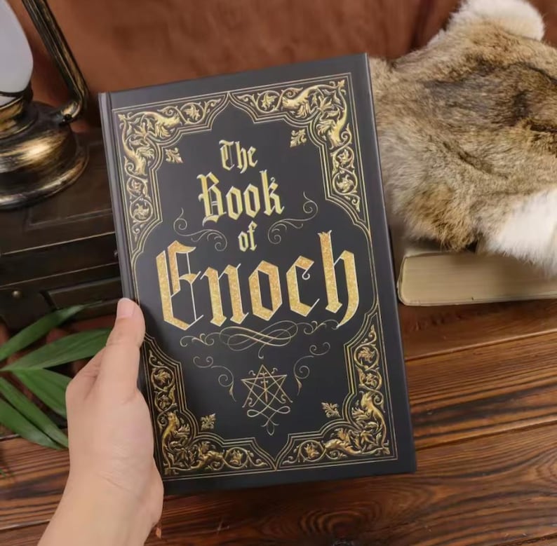 May include: A black hardcover book titled The Book of Enoch with gold foil lettering and intricate gold filigree border designs on the front cover. A small geometric symbol is printed in gold below the title text on the matte black surface.