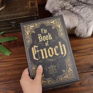 May include: A black hardcover book titled The Book of Enoch with gold foil lettering and intricate gold filigree border designs on the front cover. The cover features a central star-like symbol below the title text, all printed in a metallic gold.