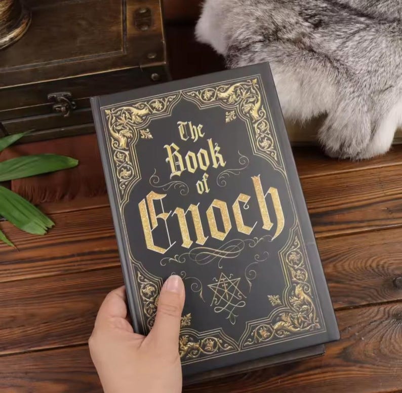 May include: A black hardcover book titled The Book of Enoch with gold foil lettering and intricate gold filigree border designs on the front cover. The cover features a central star-like symbol below the title text, all printed in a metallic gold.