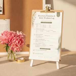 May include: A Recovery Patterns & Body Wisdom Log printed on a white sheet, secured by a gold metal clip on a wooden clipboard. The clipboard rests on a small white easel. Beside it sits a clear glass vase containing pink peony flowers.