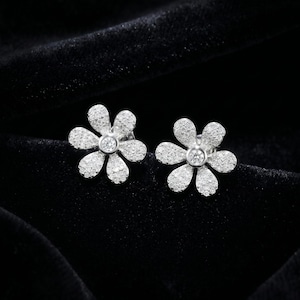 May include: A pair of silver-toned stud earrings shaped like six-petaled flowers. Each petal is covered in small, sparkling clear stones, and a single, larger round clear stone is set in the center of each flower-shaped earring.