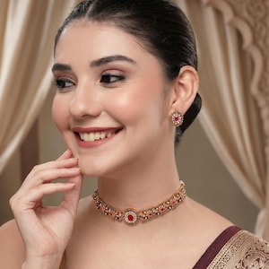 Puede incluir: Un collar tipo gargantilla en tono dorado con una piedra roja ovalada central rodeada de pequeños detalles blancos y metal labrado. Los pendientes circulares a juego con una piedra roja central y borde blanco completan el conjunto.