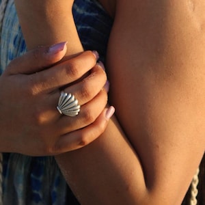 May include: A silver-toned ring shaped like a seashell with distinct vertical ridges. The ring is worn on the ring finger of a hand, featuring a metallic finish that catches the light, showcasing a detailed, fan-like scallop shell design.
