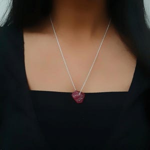 May include: A silver chain necklace with a raw, heart-shaped, deep red gemstone pendant. The pendant is a rough, natural stone with a textured surface, suspended from a delicate silver chain. The necklace is displayed against a black background.