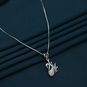 Può includere: Una collana d'argento con un ciondolo a forma di cigno. Il ciondolo presenta un design a cigno con un corpo incastonato con piccole pietre scintillanti. La catena della collana è una catena fine a scatola. Lo sfondo è un tessuto verde acqua scuro.