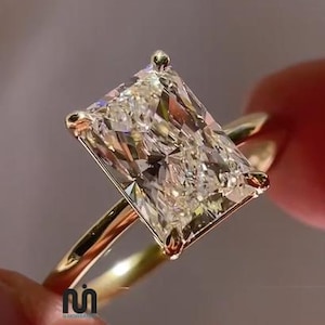 May include: A radiant-cut clear gemstone ring set in a polished yellow gold band. The rectangular stone features multiple facets that reflect light, secured by four small prongs. A small logo with the letter M is visible on the gold band.