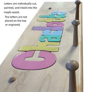 Peut inclure: Un porte-manteau en bois d'érable avec le nom Charlotte incrusté de lettres découpées et peintes individuellement en rose, jaune, bleu sarcelle et violet. Il comporte trois patères en bois. Texte sur l'image : Letters are individually cut, painted, and inlaid into the maple wood. The letters are not placed on the top or engraved.