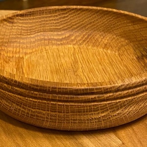 Puede incluir: Un cuenco redondo de madera con un acabado natural de vetas. El cuenco tiene un diseño ancho y poco profundo con un detalle exterior acanalado. El cuenco está hecho de madera marrón claro y se asienta sobre una superficie de madera.