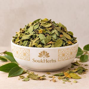 Può includere: Una ciotola in ceramica bianca riempita di foglie verdi essiccate, caratterizzata da un motivo floreale dorato e dal testo SoukHerbs stampato sul davanti. La ciotola è circondata da foglie secche sparse e alcune foglie verdi fresche.