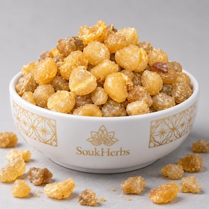 May include: A white ceramic bowl with a gold geometric pattern and the text SoukHerbs, filled with a mound of translucent, amber-coloured resin tears. Several small, irregular pieces of the same resin are scattered around the base of the bowl.