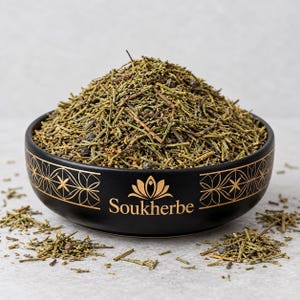 Könnte beinhalten: Eine schwarze Keramikschale mit goldenen geometrischen Mustern und dem Text Soukherbe, gefüllt mit einem Haufen getrockneter grüner Kräuterstängel und kleinen dunklen Beeren. Die Schale ist abgerundet mit matter Oberfläche und enthält holzige Pflanzenteile.