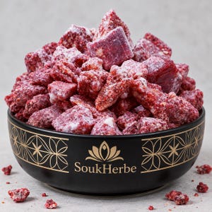 Può includere: Una ciotola in ceramica nera con motivi geometrici dorati e la scritta SoukHerbe, riempita con un mucchio di pezzi irregolari di frutta liofilizzata rosso scuro, ricoperti da una fine polvere bianca. Alcuni piccoli pezzi sono sparsi alla base.