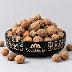 Könnte beinhalten: Eine schwarze Keramikschale mit goldenen geometrischen Mustern und dem Text SoukHerbe, gefüllt mit einem Haufen runder, strukturierter, hellbrauner getrockneter Kräuter. Einige einzelne getrocknete Kräuter liegen um den Schalenboden.