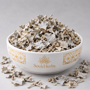 May include: A white ceramic bowl filled with dried, pale greyish-green jagged leaves and small stems. The bowl features a gold geometric pattern on the sides and the text SoukHerbs printed in gold. Scattered dried leaves surround the base of the bowl.