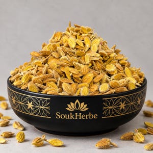 May include: A black bowl with gold geometric patterns and the text SoukHerbe, filled with a large mound of dried, golden-brown, ribbed seeds. The seeds have a textured, fuzzy appearance and are scattered around the base of the bowl.