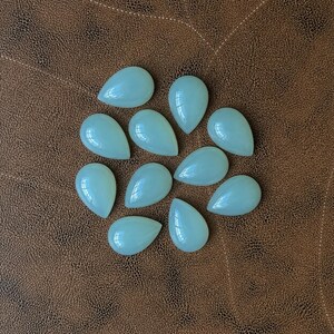 Pu&ograve; includere: Una collezione di undici gemme a forma di goccia di colore verde pallido. Le pietre lucide sono disposte su una superficie marrone simile alla pelle. Ogni pietra ha una finitura liscia e lucida, che riflette la luce. Le gemme sono probabilmente per la creazione di gioielli.