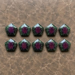 May include: Ten pentagonal faceted gemstones arranged in two rows of five. Each stone features a vibrant magenta center surrounded by a speckled green and black border, creating a distinct contrast within the geometric shape of the polished surface.