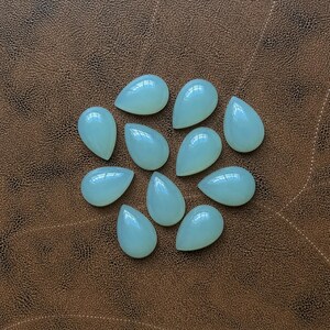 Pu&ograve; includere: Una collezione di dieci gemme a forma di goccia di colore verde pallido. Le pietre lucide sono disposte su una superficie marrone strutturata, ognuna con una parte superiore liscia e arrotondata e una base appuntita. Le gemme sono traslucide.